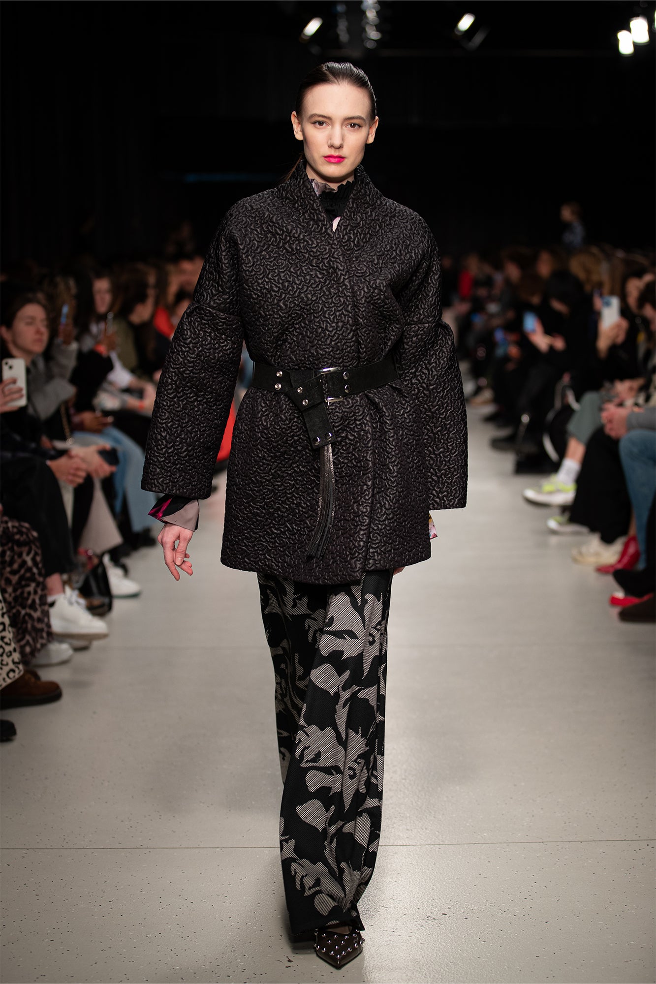 Model walking on a runway wearing a black coat and patterned pants.