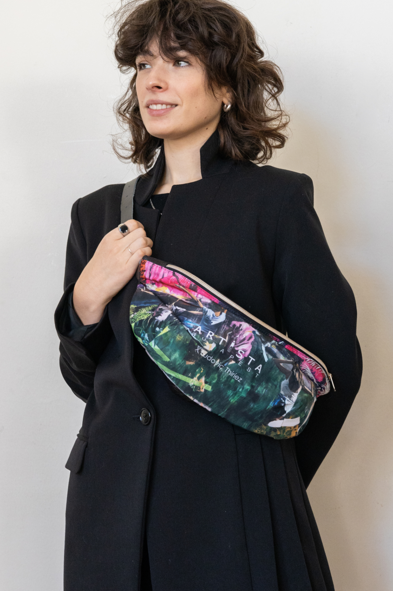 A person wearing a black coat and a multicolored floral bum bag with an adjustable leather strap.