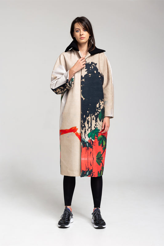 A model wearing a knee-length coat with an asymmetric print, featuring a black collar, beige body, and colorful floral and abstract patterns. The coat has a button-down closure and side pockets.