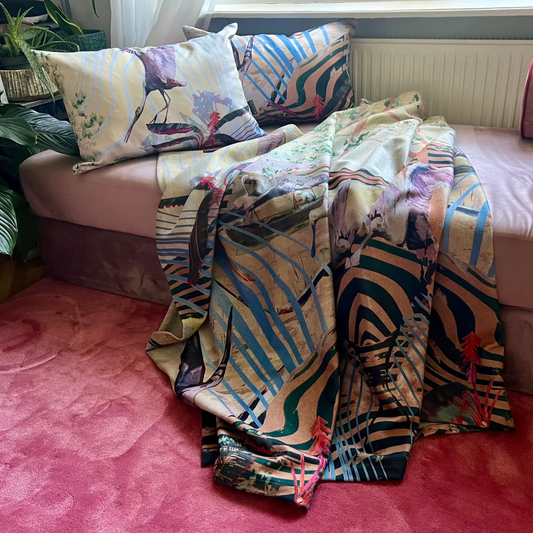 A colorful patterned throw blanket and matching pillows with tassels are draped over a pink daybed in a home setting with a pink rug and plants.