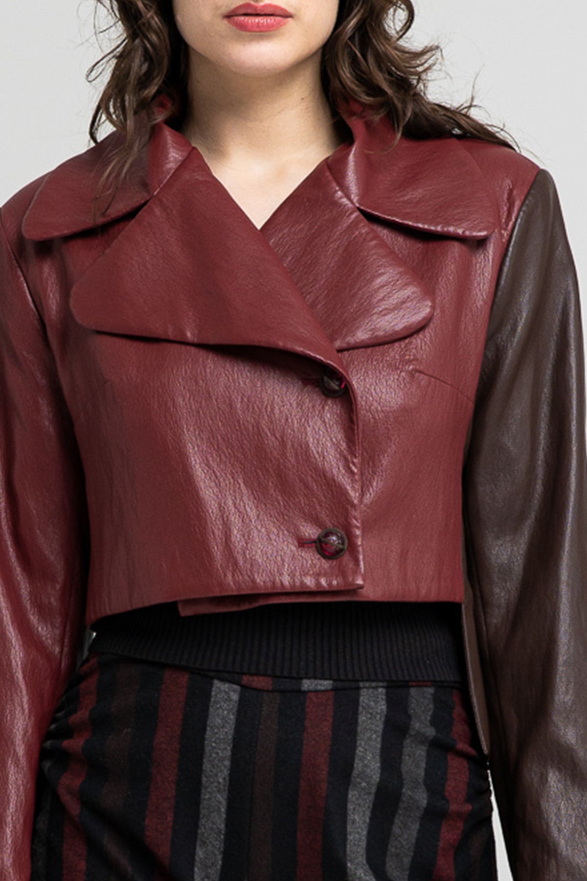 A close-up of a model wearing a cropped eco-leather jacket with a deep burgundy red body and contrasting dark brown sleeves, finished with reverse V-neckline.