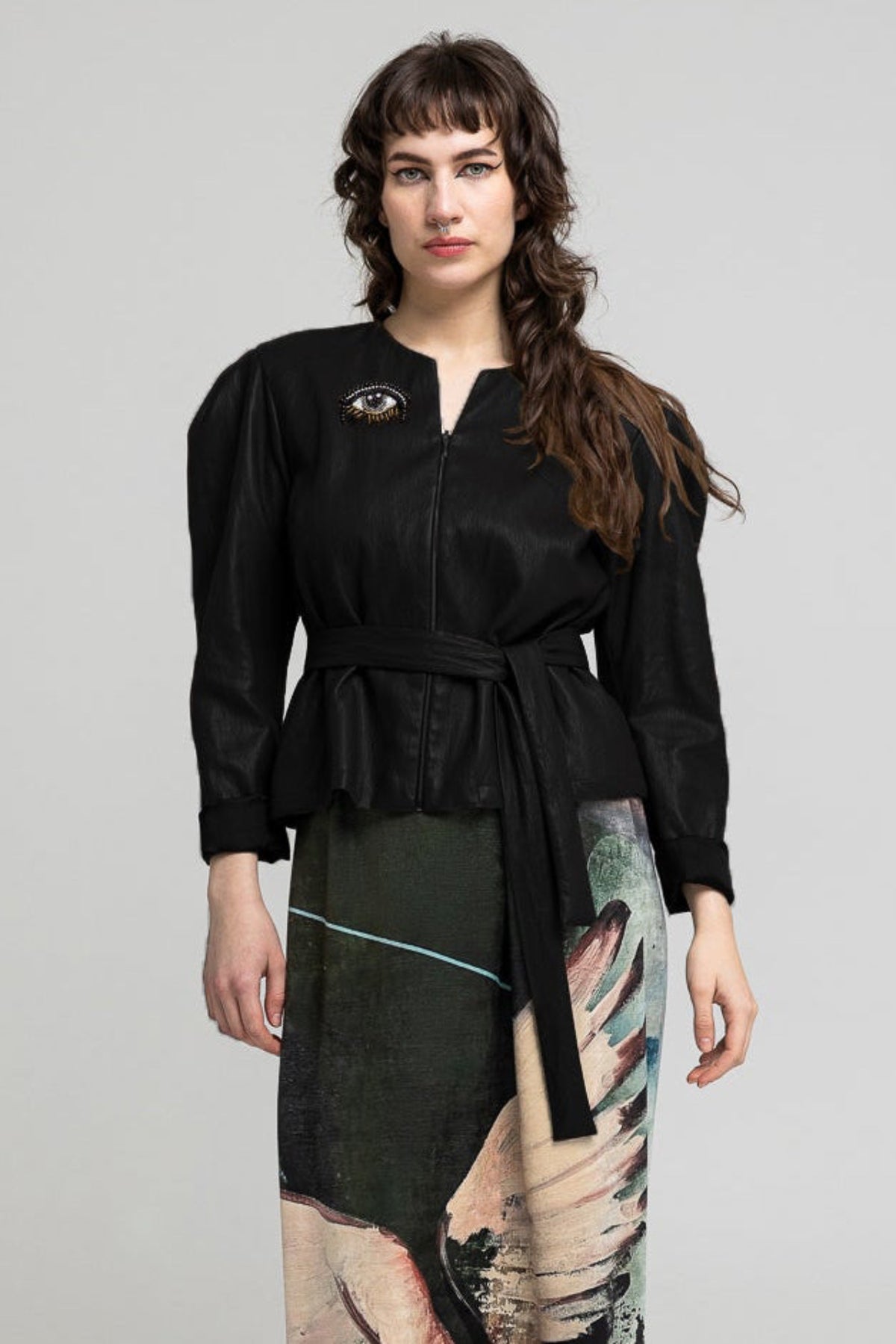 A woman stands against a light gray studio background wearing a long black belted top and a long printed skirt featuring a winged figure in dark green, beige, pink, and purple tones. She is wearing metallic brown ankle boots.