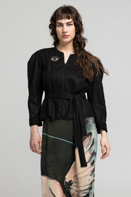 A woman stands against a light gray studio background wearing a long black belted top and a long printed skirt featuring a winged figure in dark green, beige, pink, and purple tones. She is wearing metallic brown ankle boots.