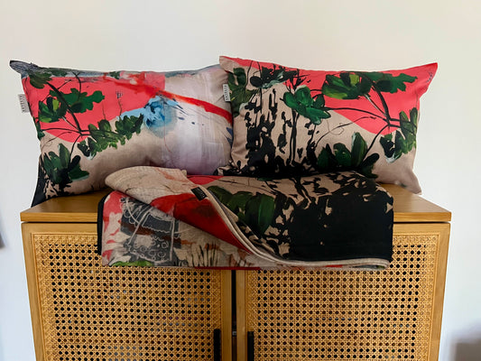 Three decorative throw pillows and a folded matching blanket with an abstract botanical print in coral pink, green, black, and beige, arranged on a wooden cabinet with a woven cane front against a plain white wall.