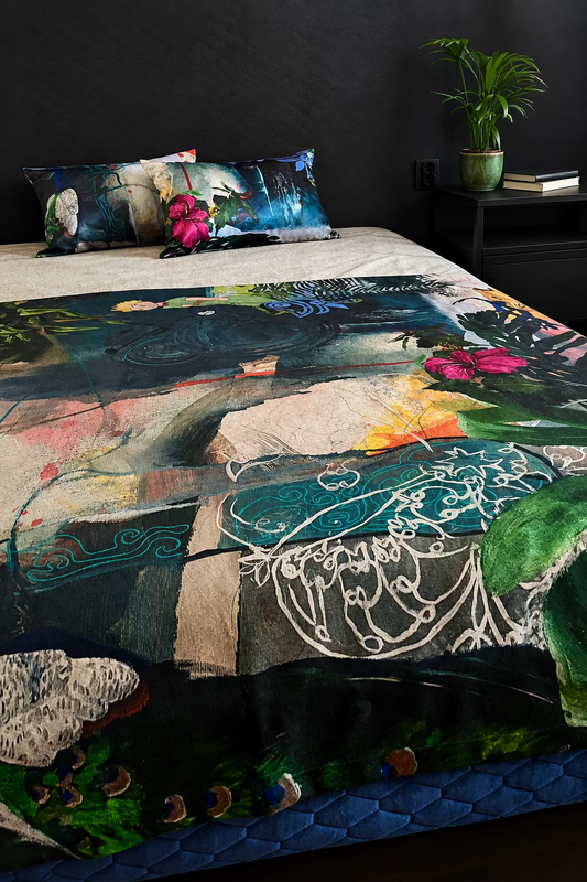 A bed dressed in a large abstract art print blanket with a matching  pillows, styled in a bedroom setting with a side table and plant.