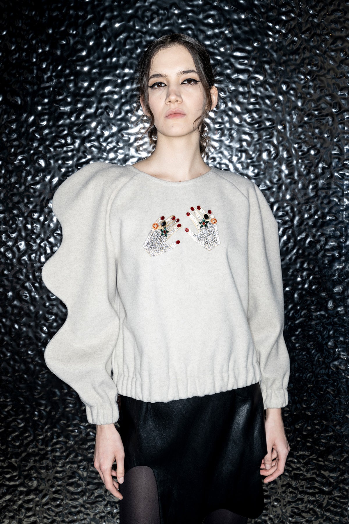 A model wearing a light beige knit sweater with dramatic wavy balloon sleeves and embroidered details on the chest, paired with a black skirt, set against a dark textured background.