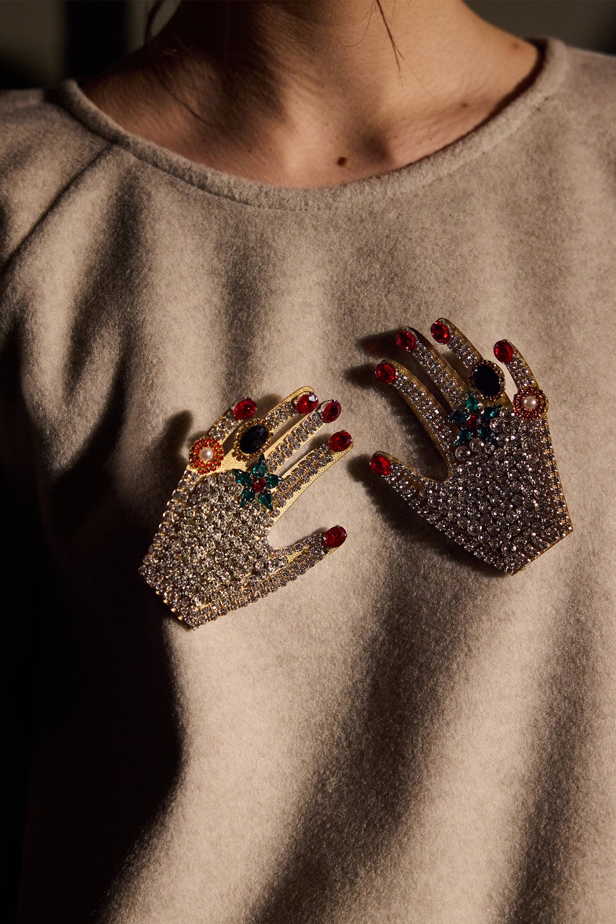 A close-up of a person wearing a beige top, featuring two hand-shaped brooches embellished with rhinestones and colorful gemstones on the chest.