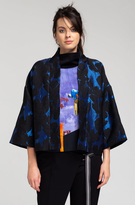 A person standing wearing a midnight blue jacket with a geometric pattern, black trousers, and black sneakers.