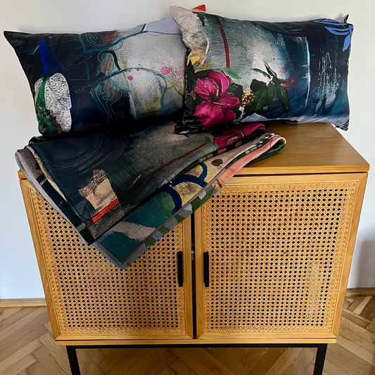 Two rectangular decorative pillows and a folded matching blanket with an abstract floral print are arranged on a light wood cabinet with woven cane doors, against a plain white wall and wood floor.