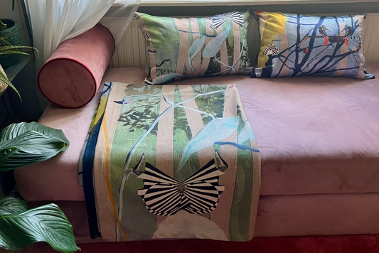 A pink upholstered bench styled with two rectangular patterned pillows and a matching folded throw blanket featuring a zebra and tropical bird motifs. A pink cylindrical bolster pillow sits on the left, with sheer white curtains and potted plants visible in a bright indoor setting.