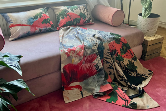 A pink sofa in a bright living room with a draped throw ablanket and matching pillows featuring  bold tropical floral print in red, green, and beige. A pink rug covers the floor, with potted plants and wooden crates nearby.