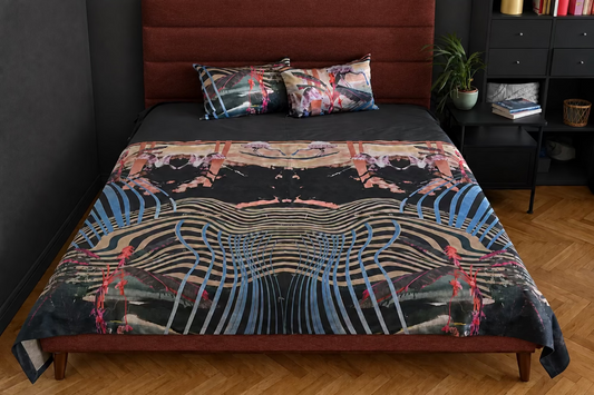 A bed dressed in a dark blue quilted headboard and matching blue bed skirt, with a bold abstract print blanket featuring black, blue, pink, and beige wavy patterns. Two matching decorative pillows rest against the headboard in a bedroom setting with a wood floor, bedside table, and white cabinet.