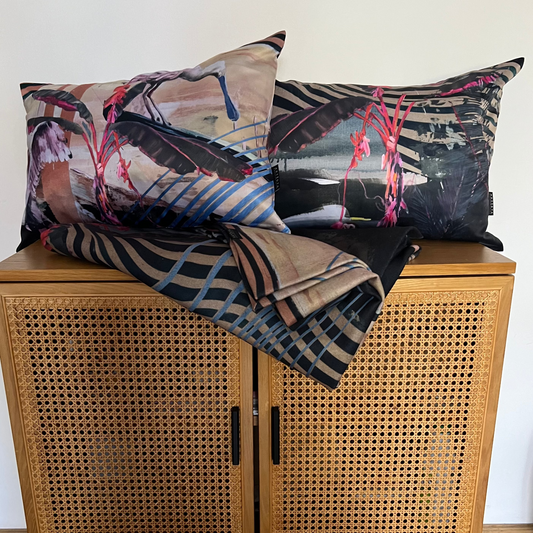 A set of two decorative throw pillows and a matching folded throw blanket with an abstract multicolor print, styled on top of a wooden cabinet with a cane front.