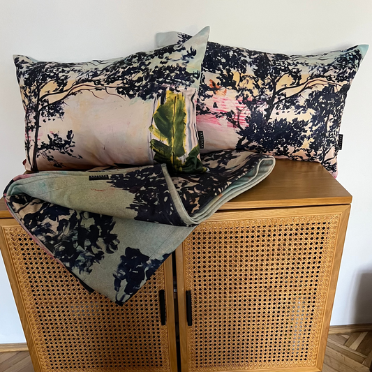 A styled home decor arrangement featuring two floral throw pillows and a matching folded blanket with a botanical leaf print, displayed on top of a wooden cane cabinet in a room with a herringbone wood floor.