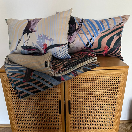A styled home decor arrangement featuring two patterned throw pillows and a folded duvet set draped over a mid-century modern wooden cabinet with cane doors, set against a plain white wall and herringbone parquet floor.